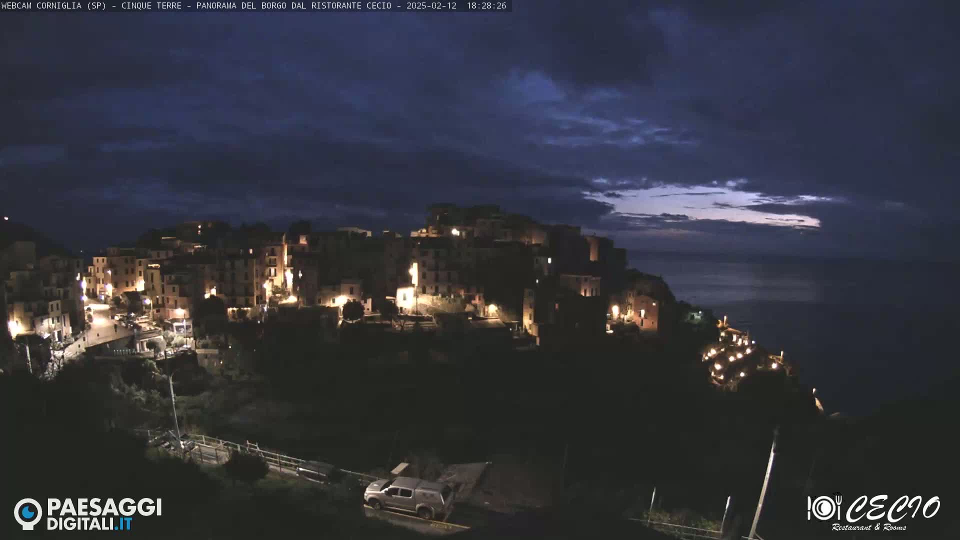 A nighttime view of a small, illuminated village perched on a cliff overlooking a dark sea under a mostly cloudy sky.
