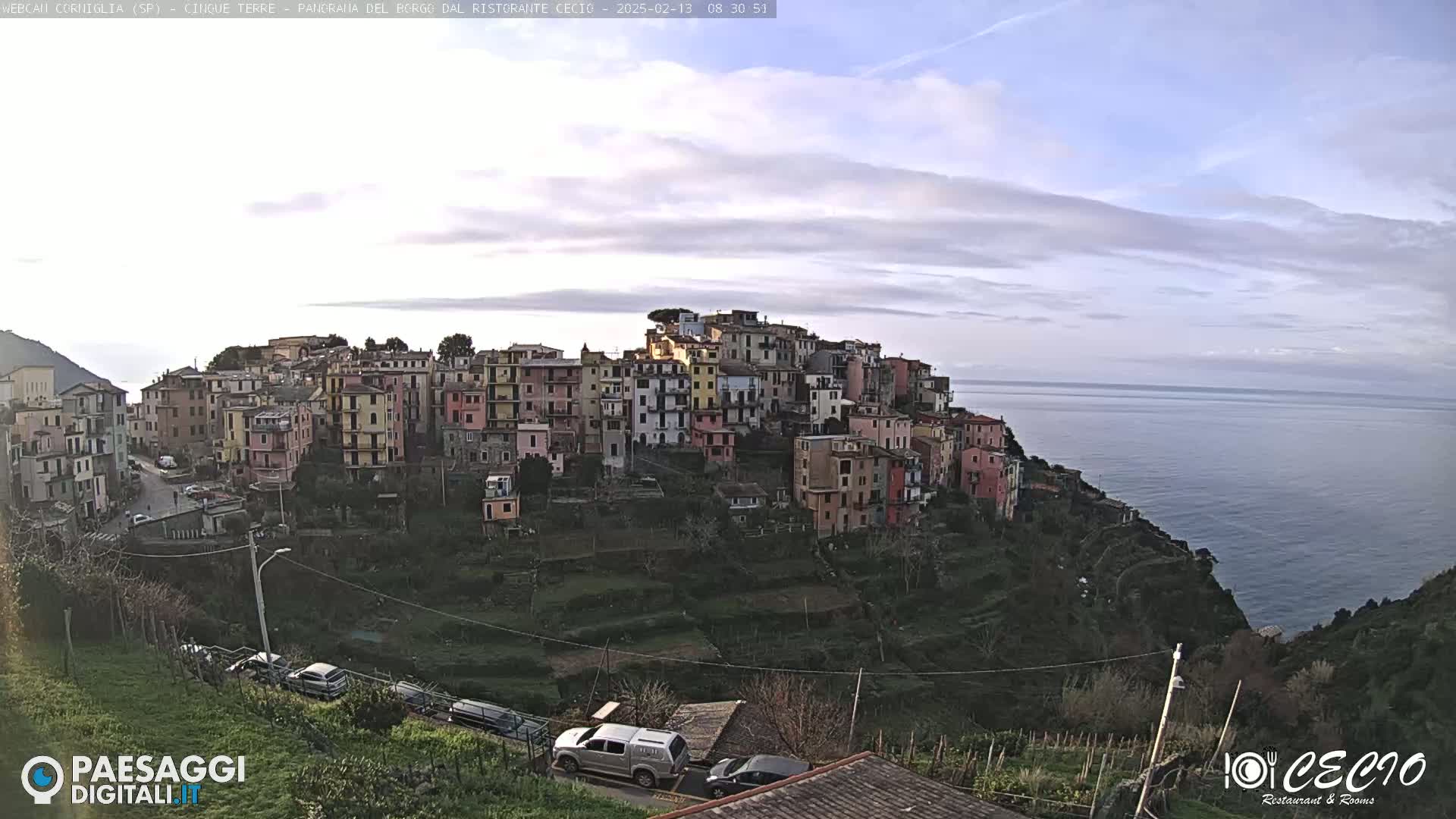 A colorful hillside village overlooks a calm ocean under a mostly cloudy sky.