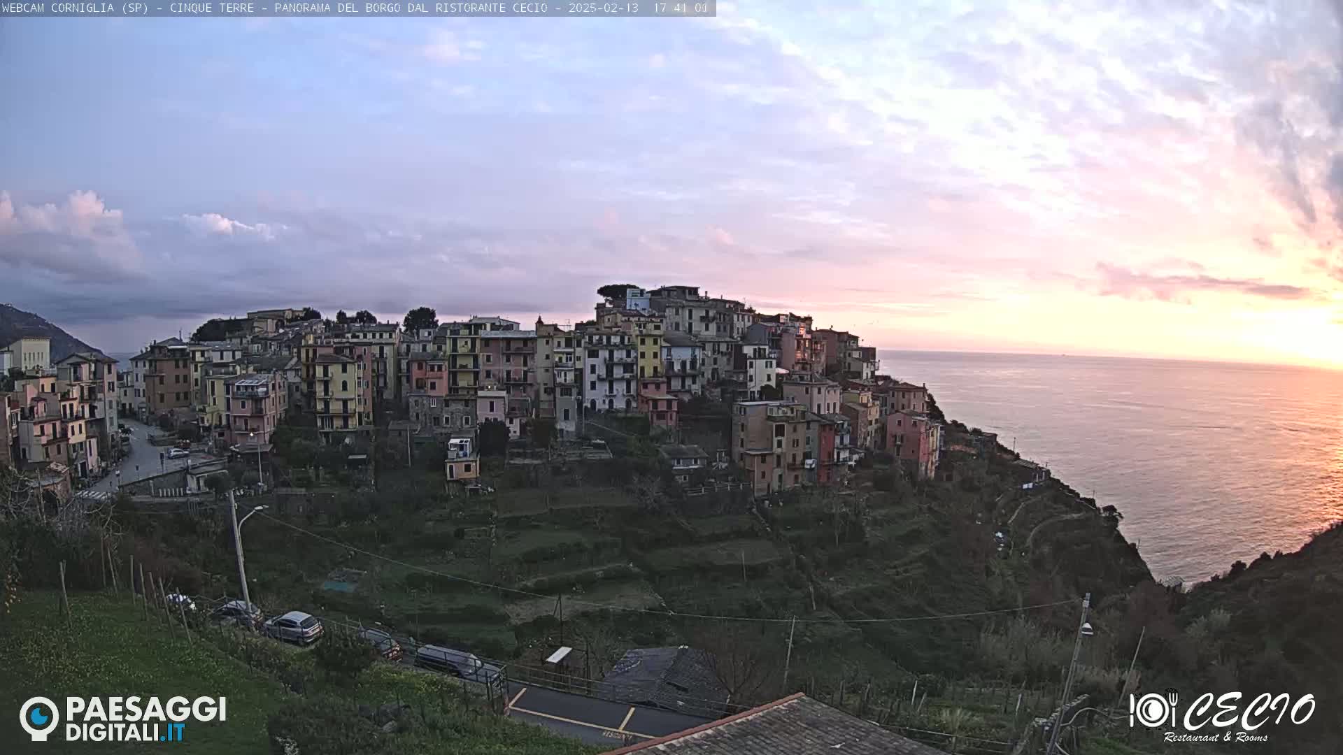 A colorful village clings to a hillside overlooking a calm sea at sunset.