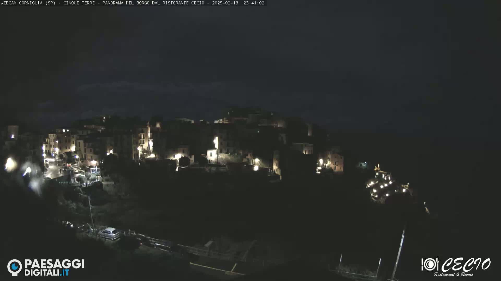 A nighttime, long shot of a small, illuminated town nestled on a hill under a dark sky.