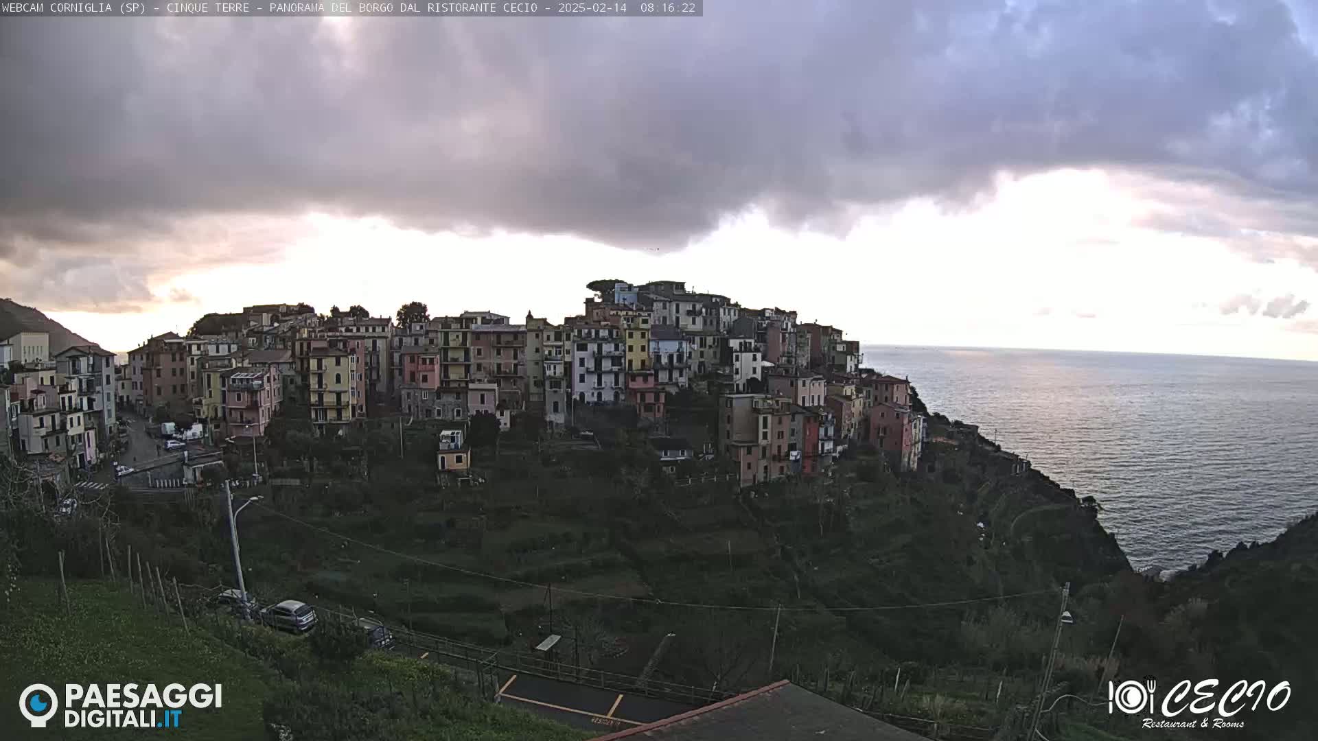 A coastal village of colorful buildings is perched on a hillside overlooking a calm sea under a mostly cloudy sky.