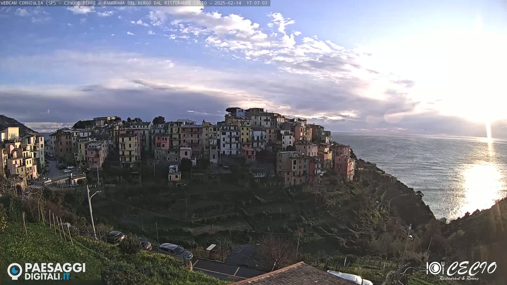 A colorful hillside village overlooks a calm sea under a partly cloudy sky.