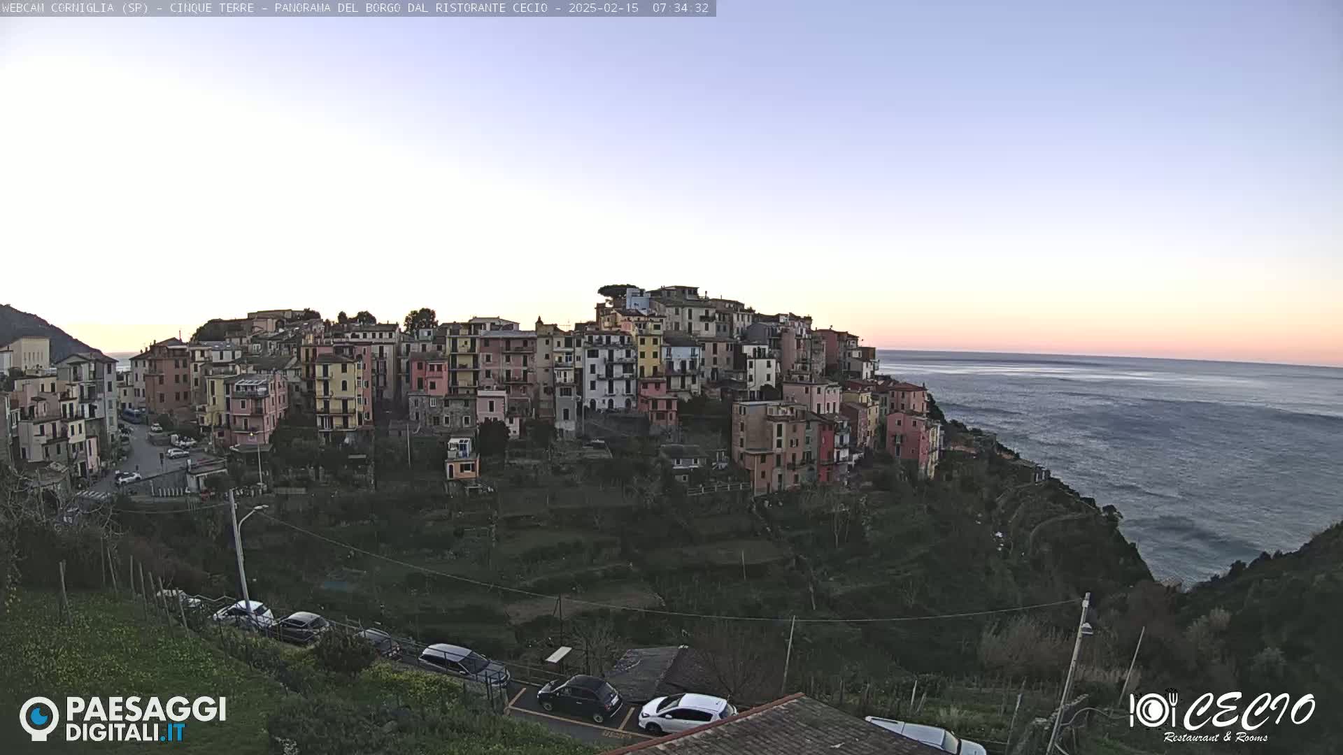 A colorful hillside village overlooks a calm ocean at dusk under a clear sky.