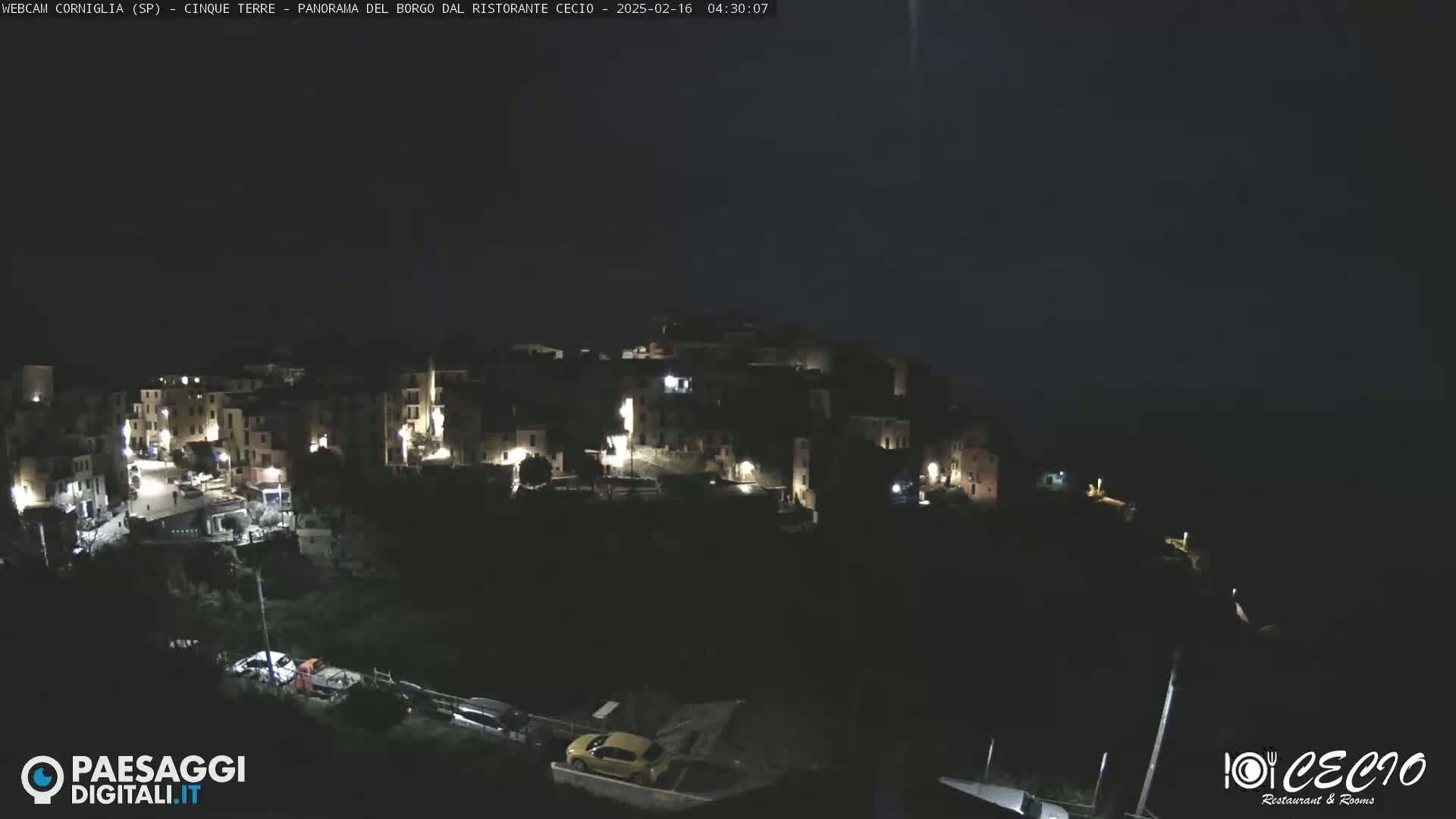 A nighttime view of a small, illuminated town nestled on a hillside overlooking a dark body of water under a mostly clear, dark sky.