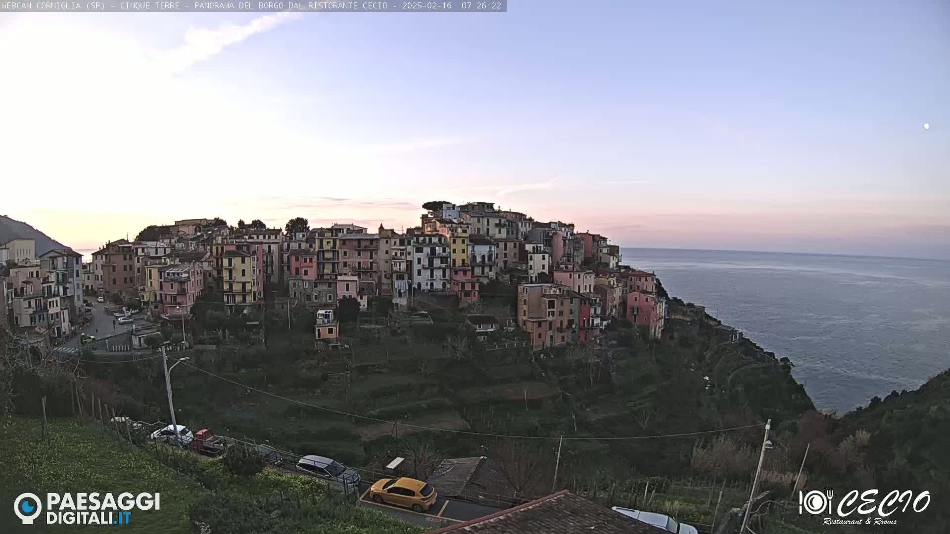 A colorful hillside village overlooks a calm sea under a pale, twilight sky.