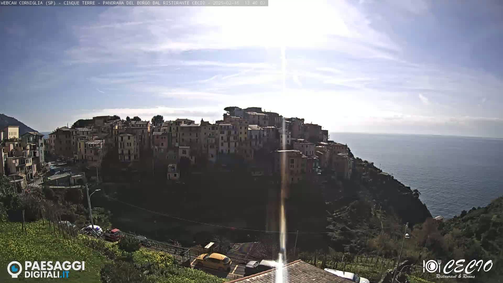 A sunlit coastal town, nestled on a hillside overlooking a calm sea under a mostly clear sky, with a few cars visible below.