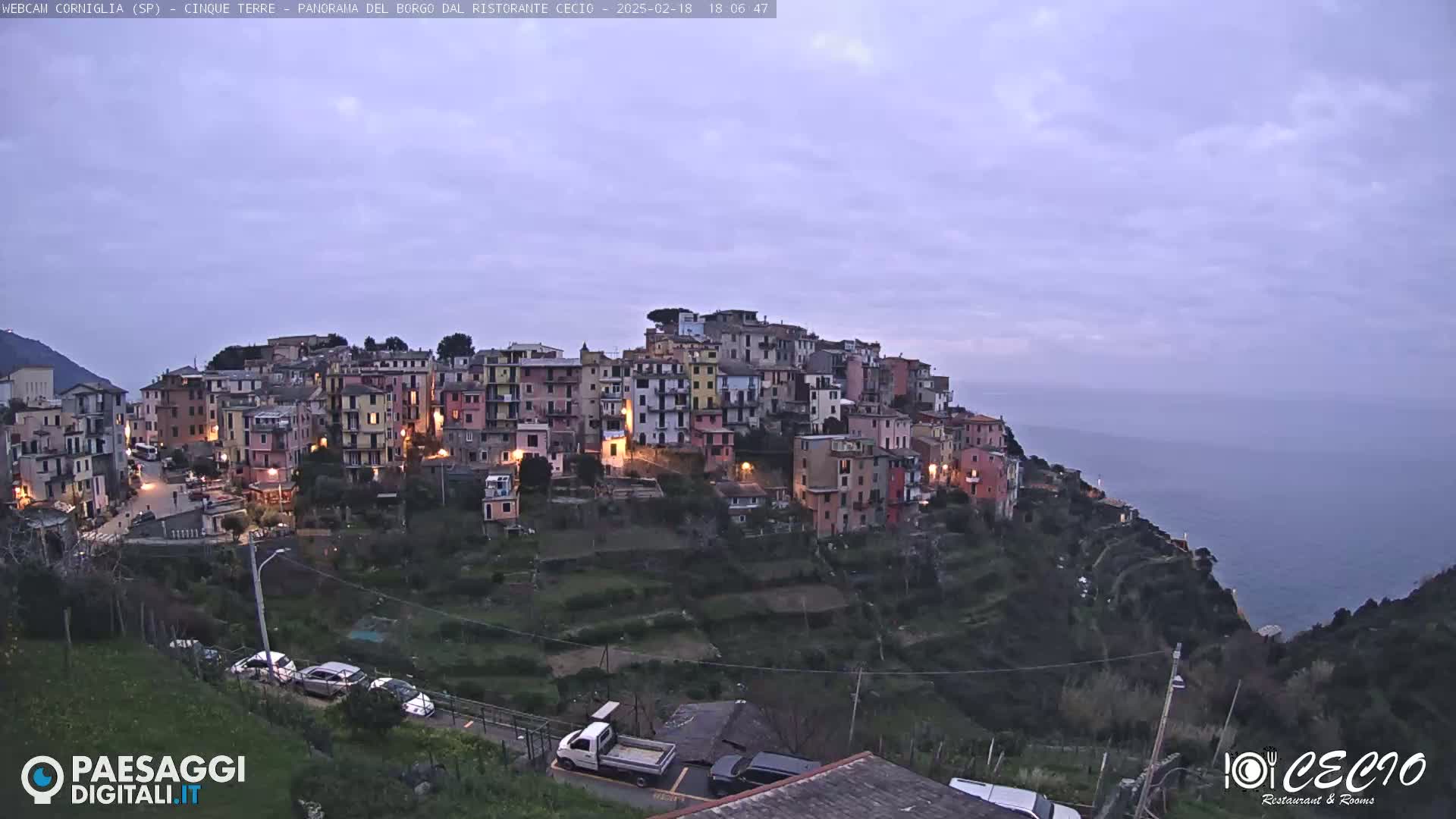 A colorful hillside village with lights on at dusk overlooks a calm sea under a pale, cloudy sky.