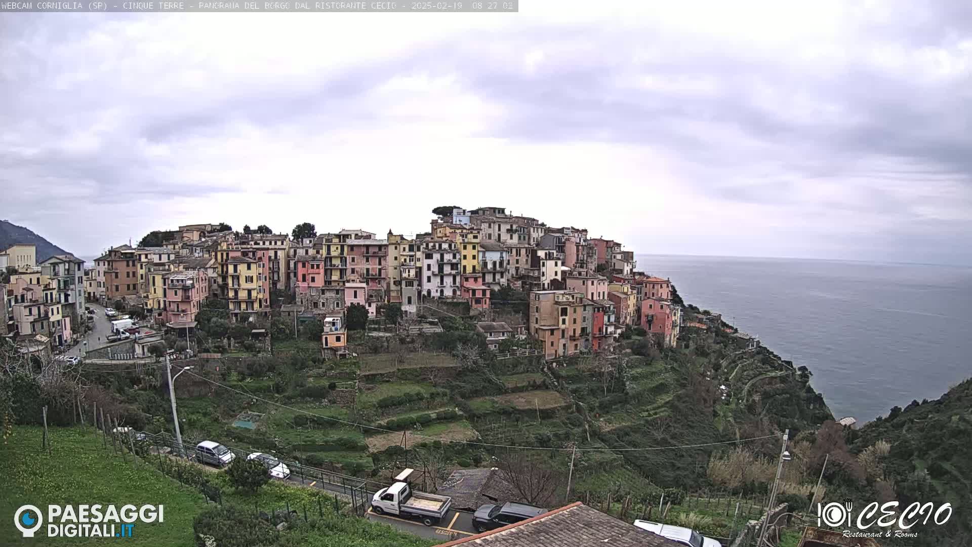 A colorful village clings to a hillside overlooking a calm sea under a cloudy sky.