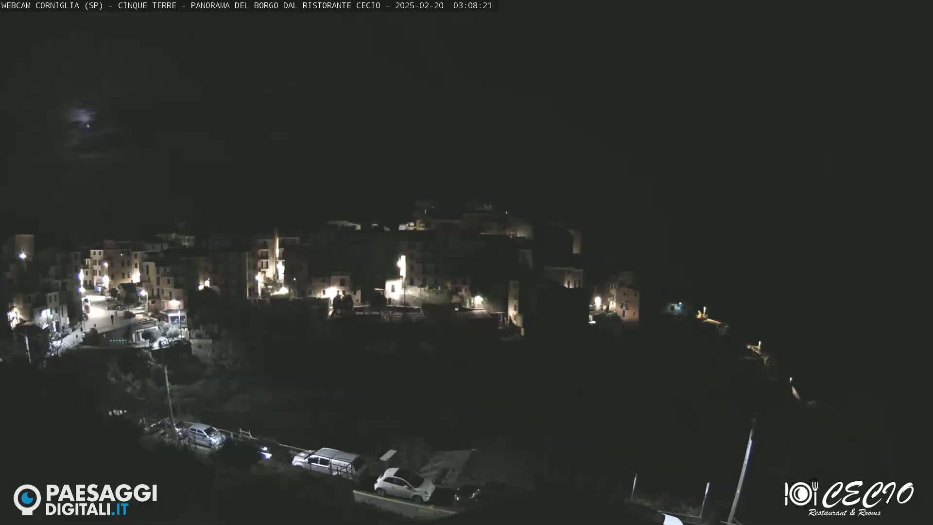 A nighttime, high-angle view shows a small, illuminated town nestled on a hillside under a dark, possibly stormy sky.