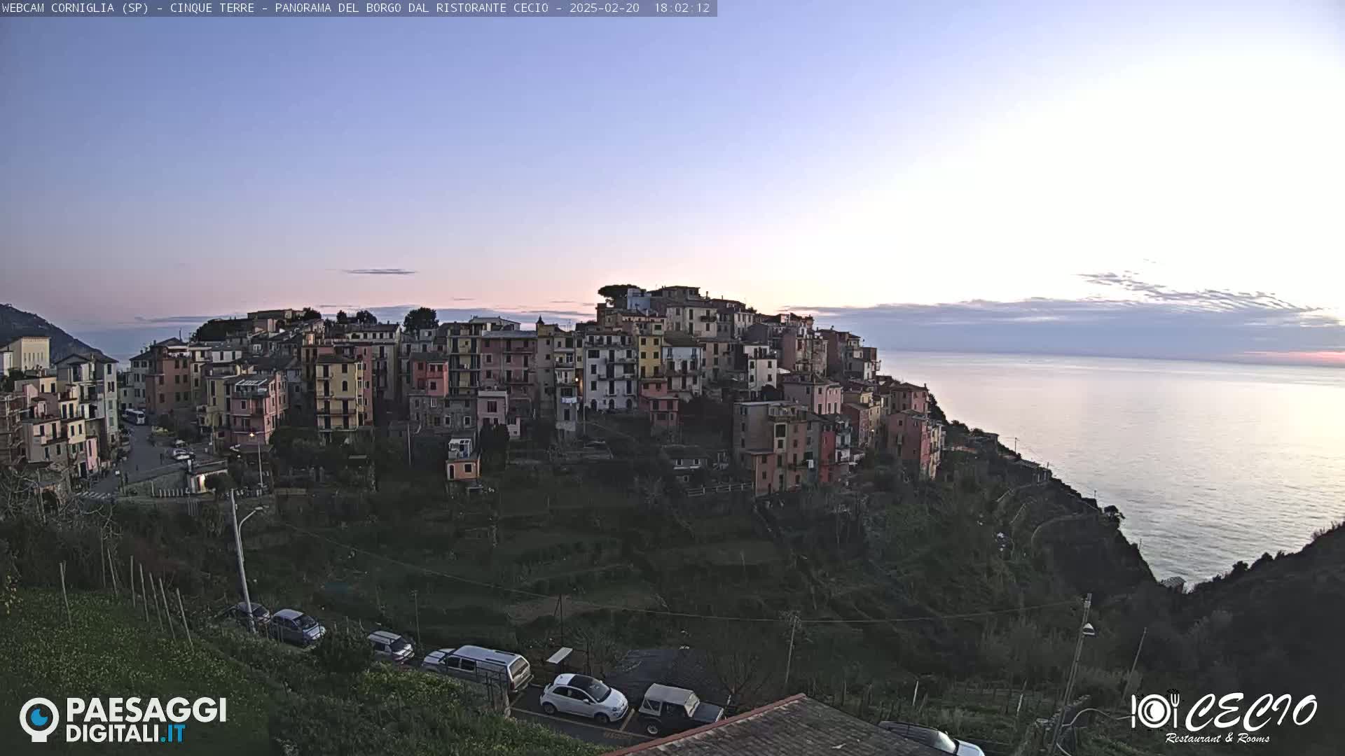 A colorful village clings to a hillside overlooking a calm sea under a twilight sky.