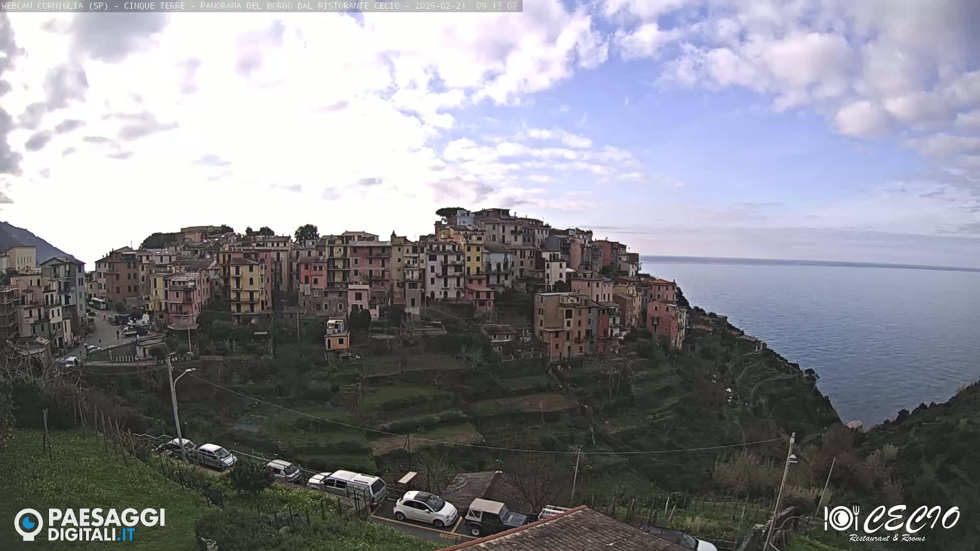 A coastal village of colorful buildings clings to a hillside overlooking a calm sea under a partly cloudy sky.