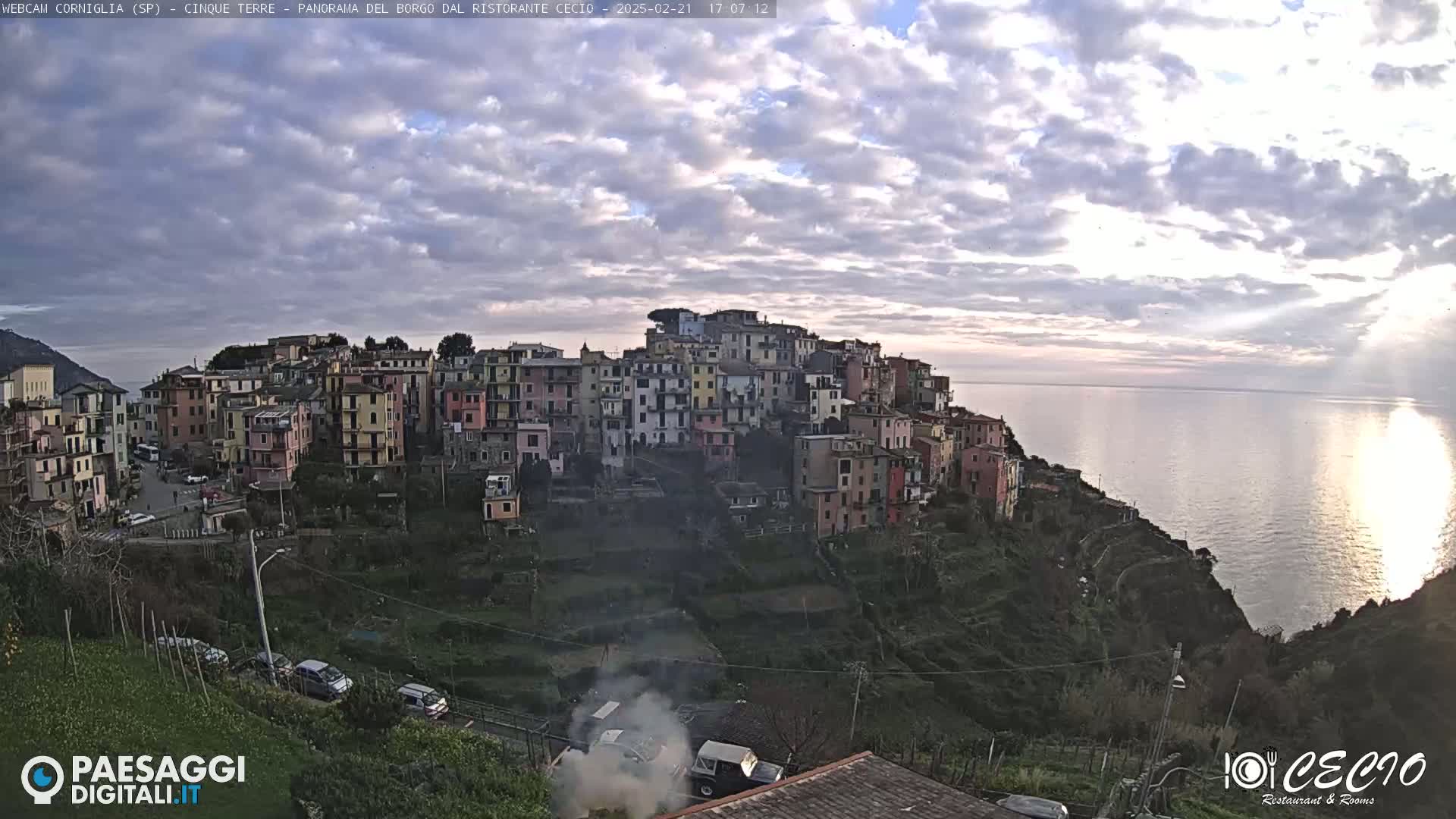 A coastal village of colorful buildings clings to a hillside overlooking a calm sea under a partly cloudy sky.