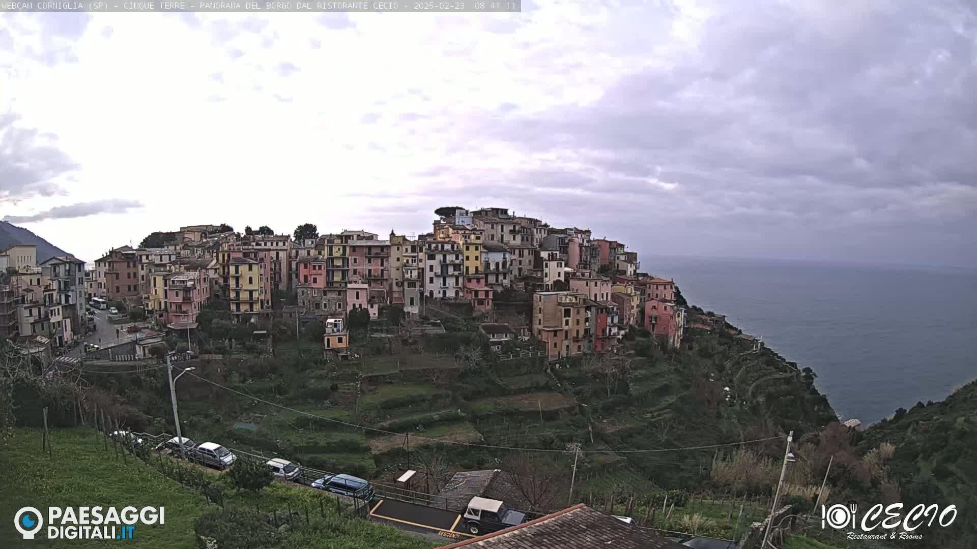 A coastal village of colorful buildings on a hillside overlooks a calm sea under an overcast sky.