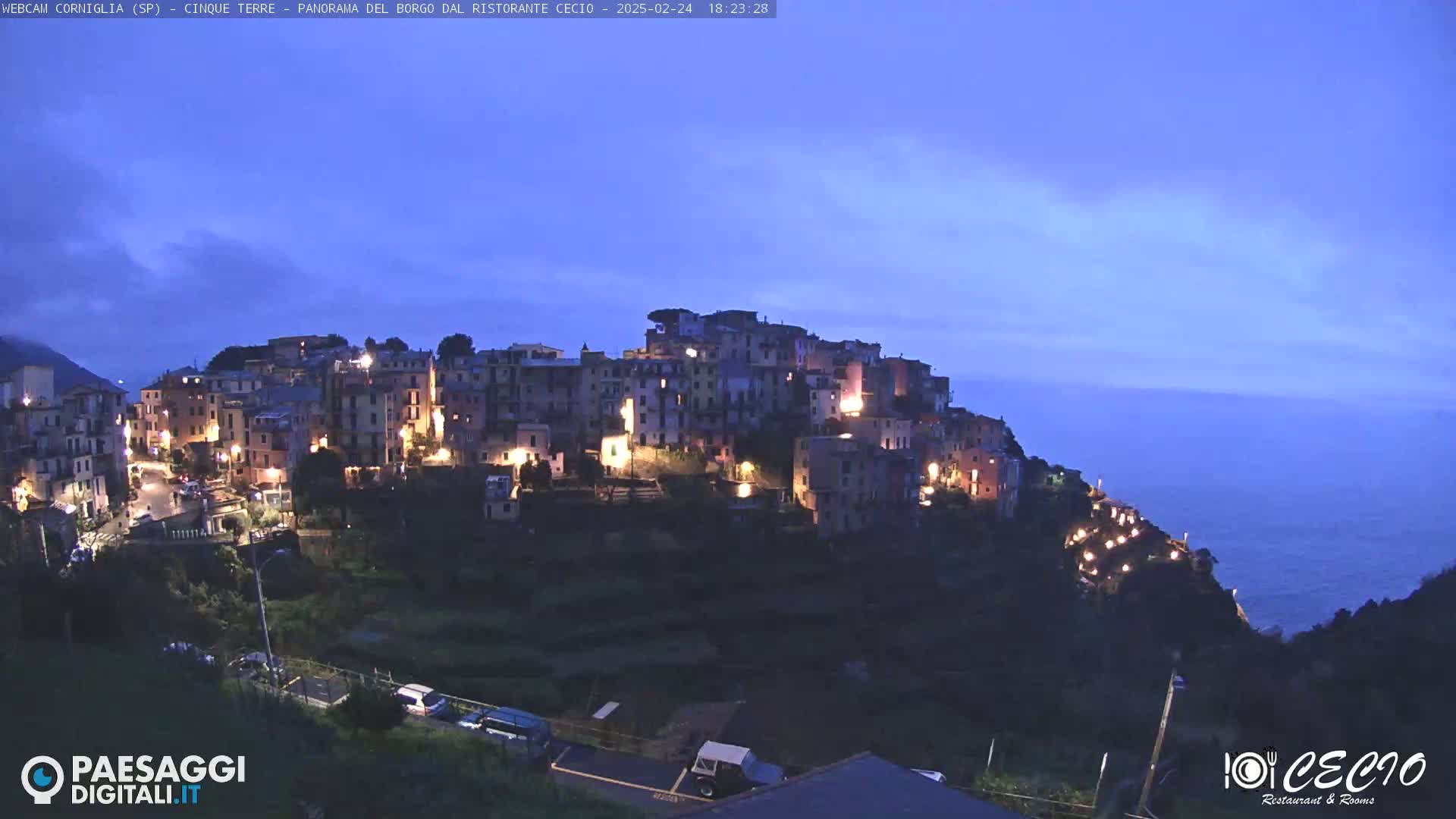 A hilltop village is illuminated at twilight, overlooking a dark blue sea under a mostly clear sky.