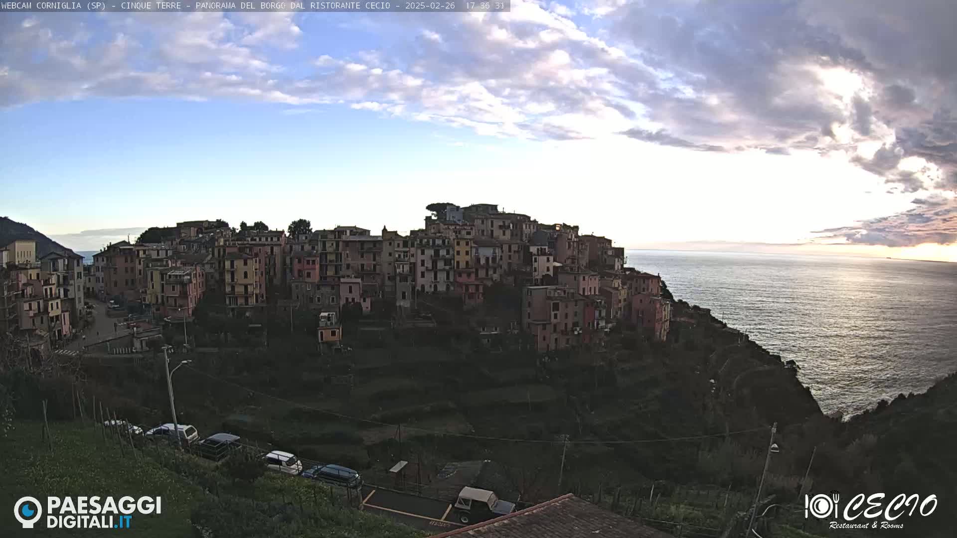 A coastal town of colorful buildings perched on a cliff overlooking a calm sea under a partly cloudy sunset sky.