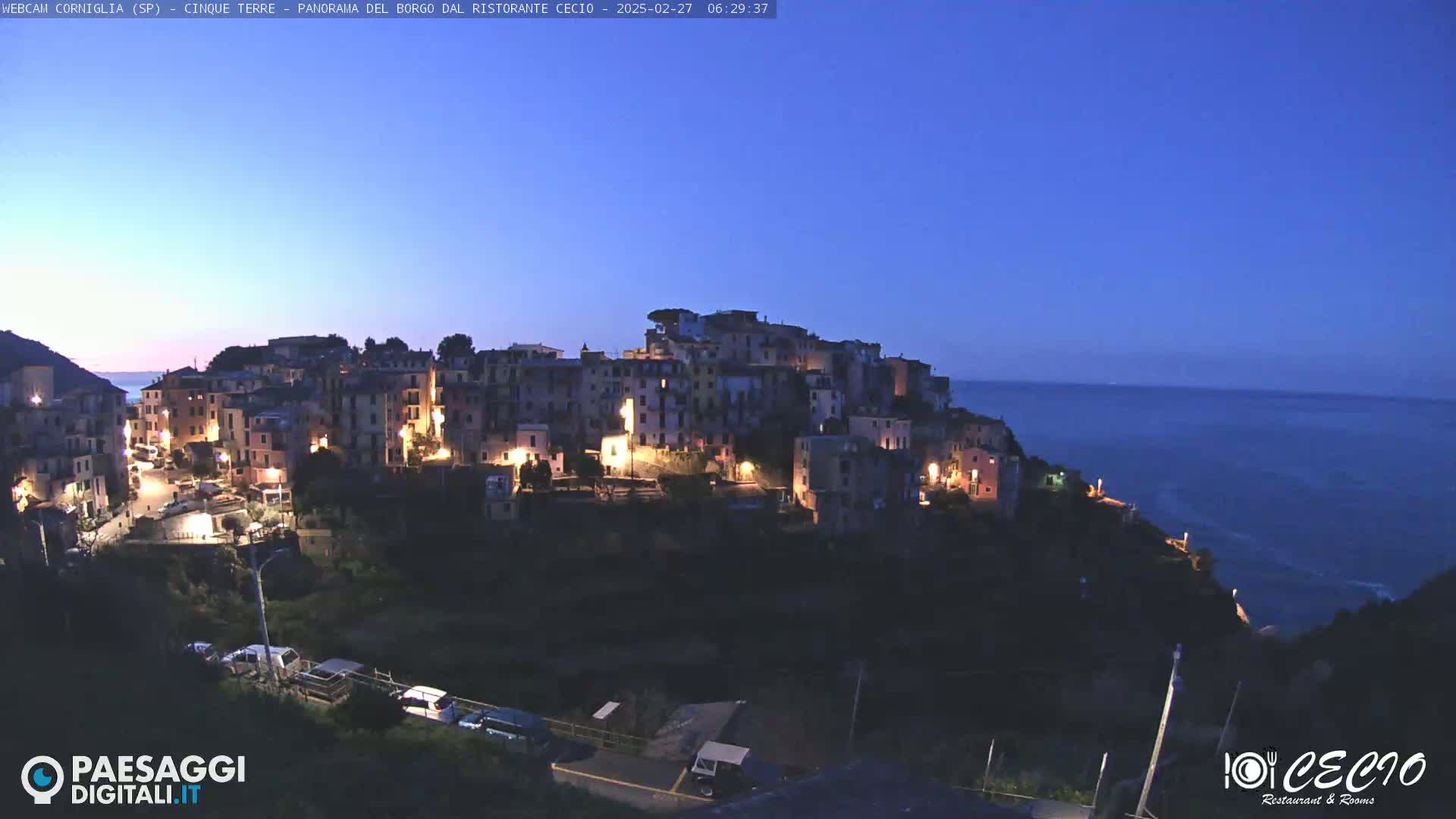 A brightly lit coastal town sits atop a dark cliff overlooking a calm, dark blue sea under a twilight sky.