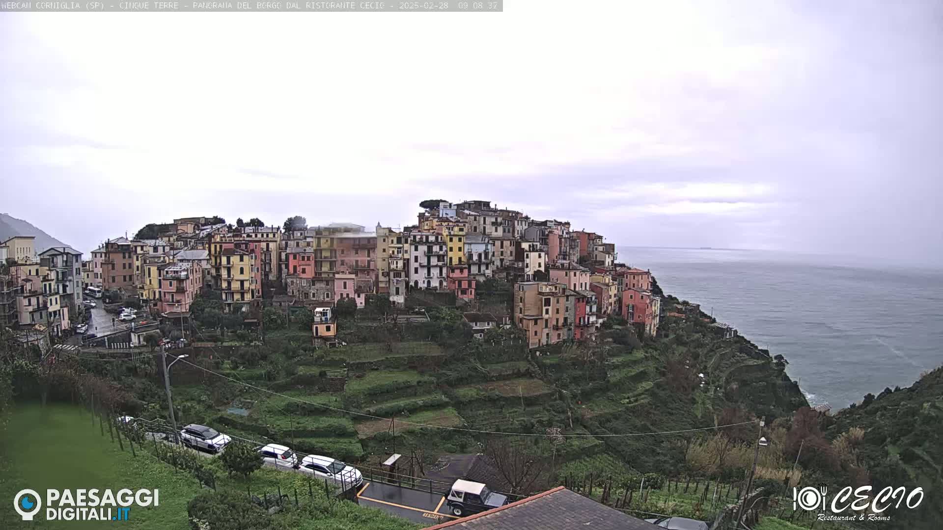 A coastal village of colorful buildings on a hillside overlooks a calm sea under an overcast sky.