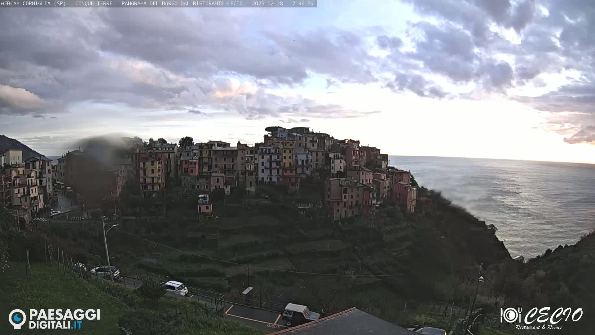 A coastal village of colorful buildings clings to a hillside overlooking a calm sea under a partly cloudy sky.