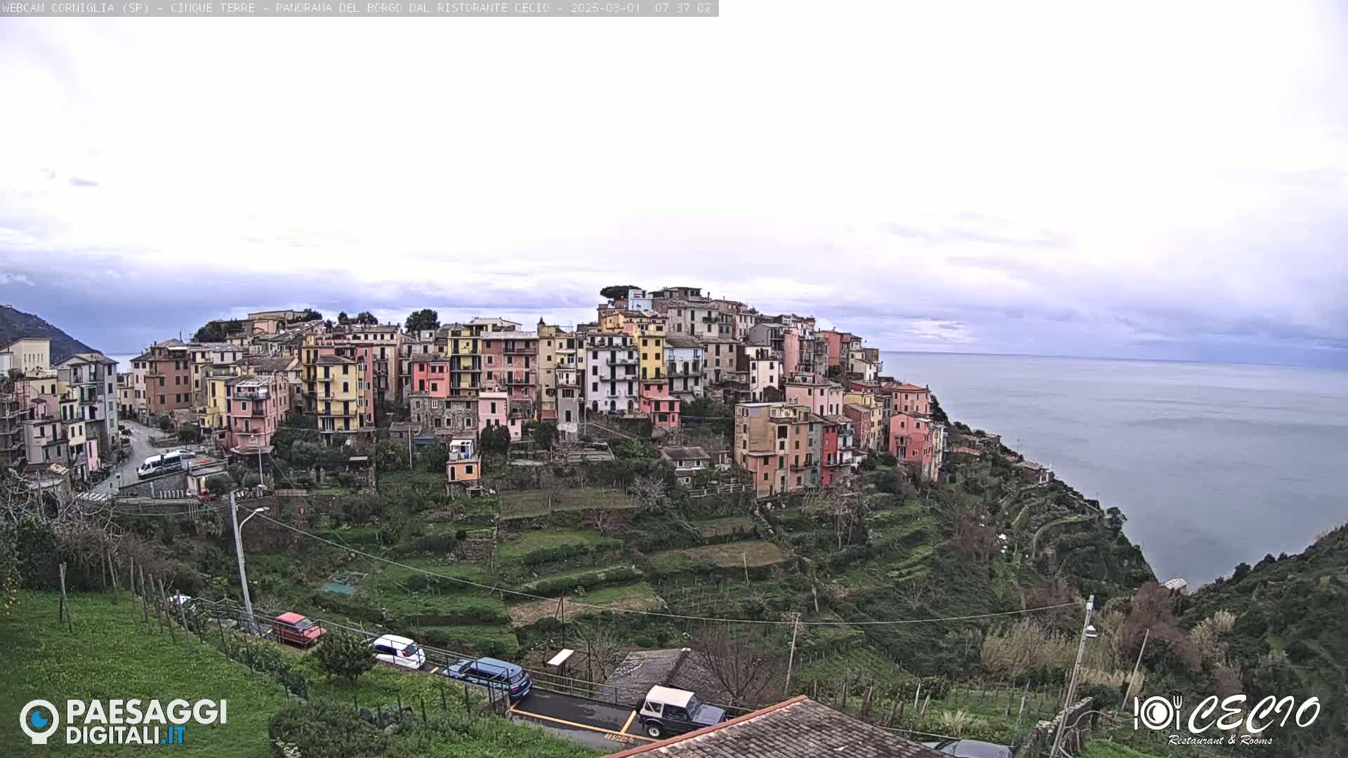 A coastal village of colorful buildings on a hillside overlooks a calm sea under an overcast sky.