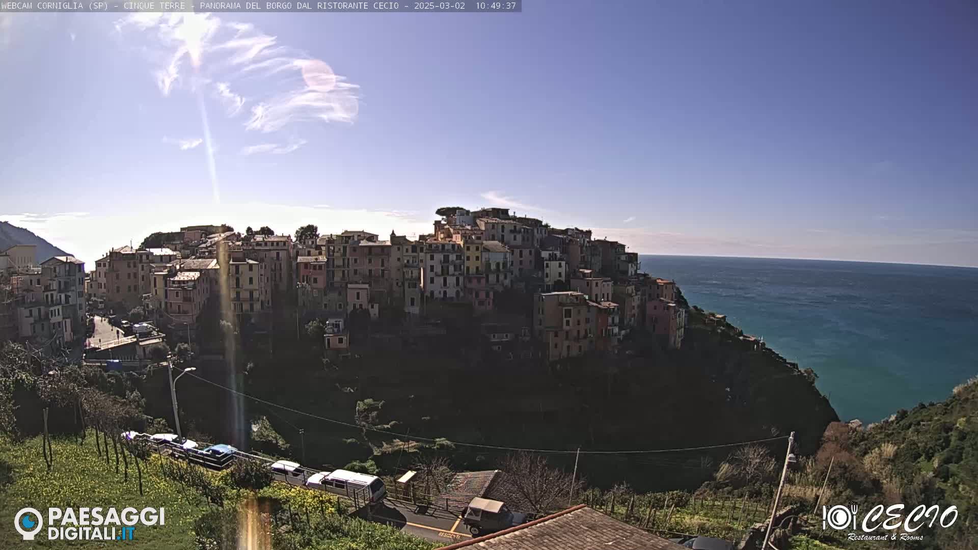A colorful hillside village overlooks a calm blue ocean under a mostly clear, sunny sky.
