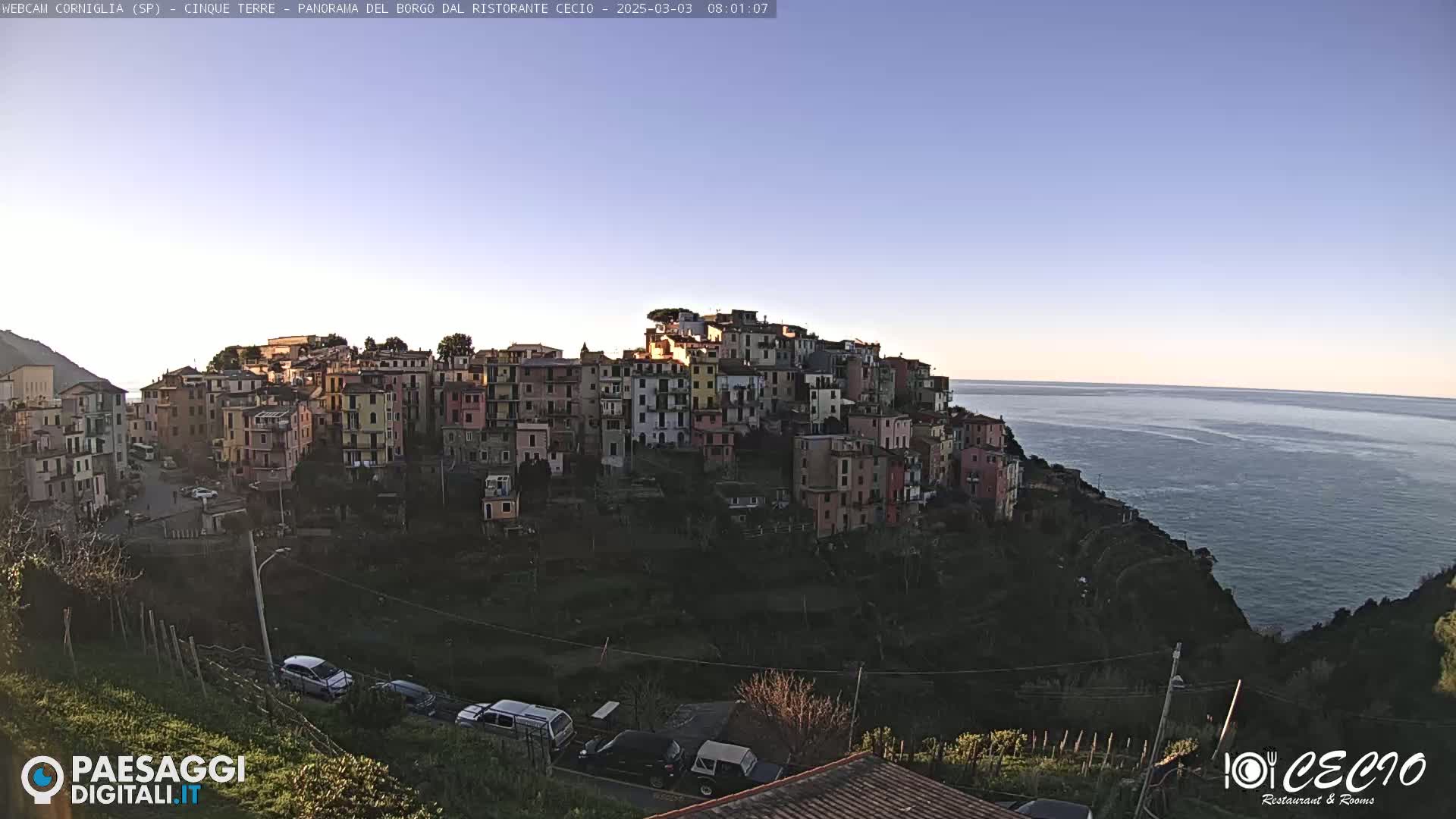 A coastal village of colorful buildings clings to a hillside overlooking a calm sea under a clear, bright sky.