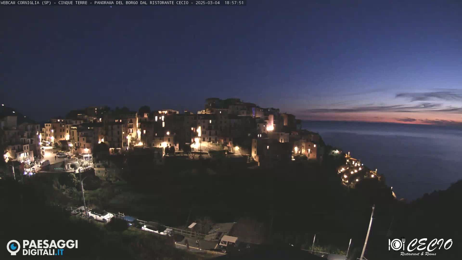 A brightly lit village clings to a dark cliffside overlooking a calm, twilight ocean.