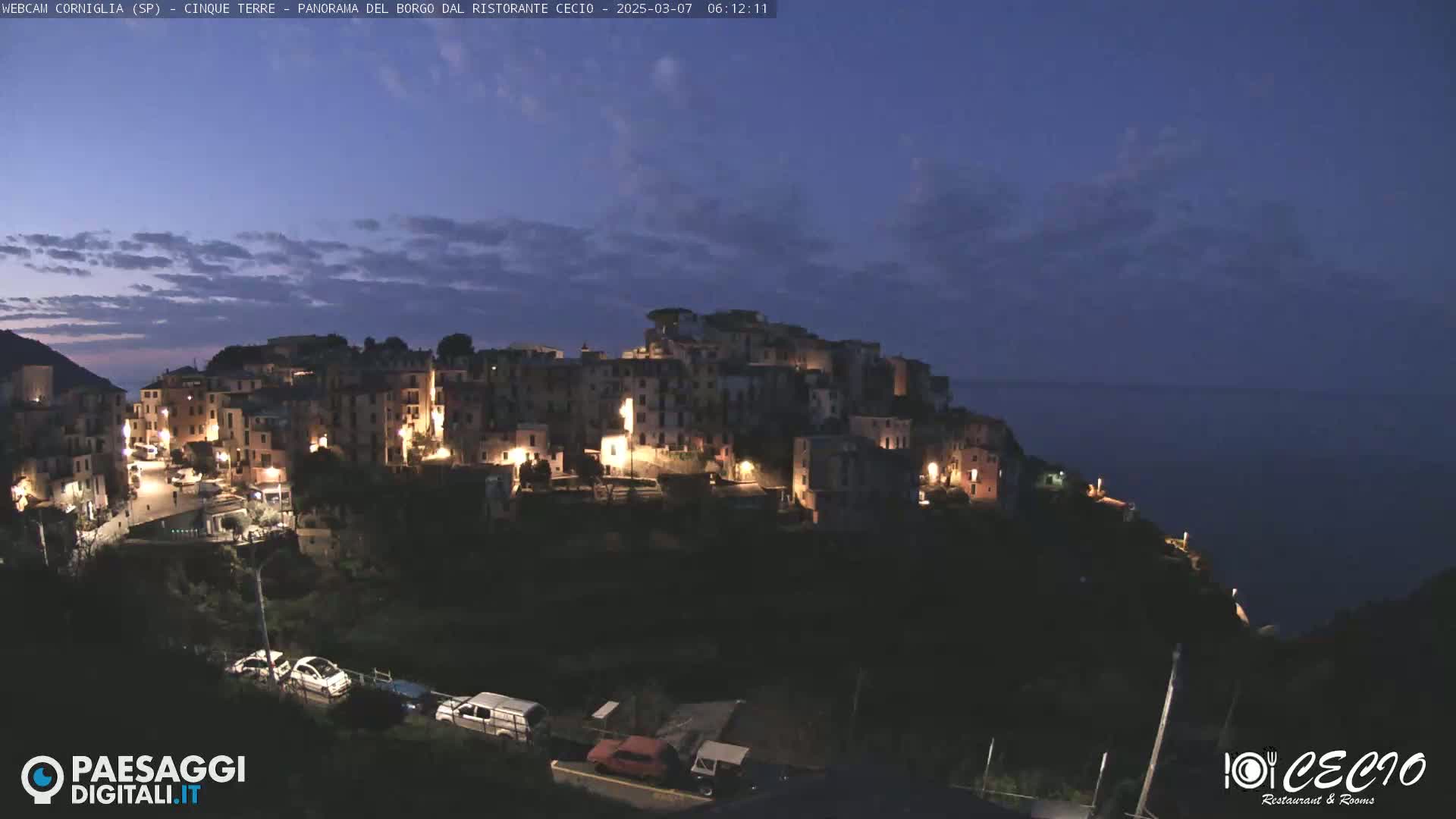 A nighttime view of a brightly lit hillside village overlooking a dark ocean under a twilight sky.