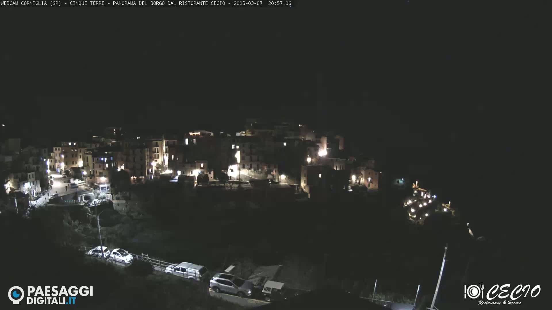 A nighttime, high-angle view shows a small village nestled on a hillside with a few cars parked below.