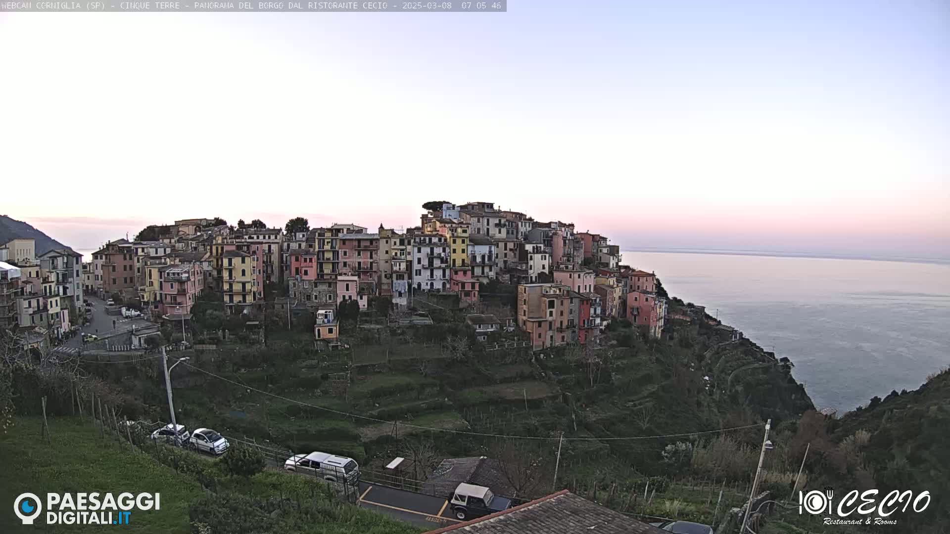 A colorful hillside village overlooks a calm sea under a pale pink and lavender sky at dawn.