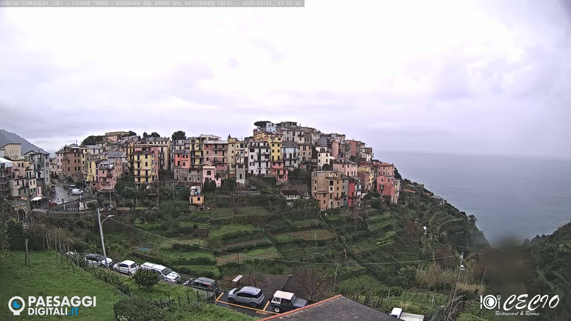 A hillside village of colorful buildings overlooks a calm ocean under an overcast sky.