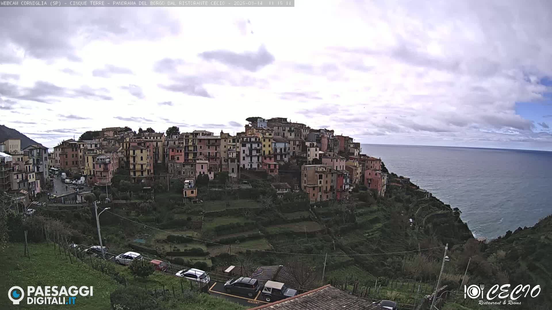 A hillside village of colorful buildings overlooks a calm ocean under a mostly cloudy sky.