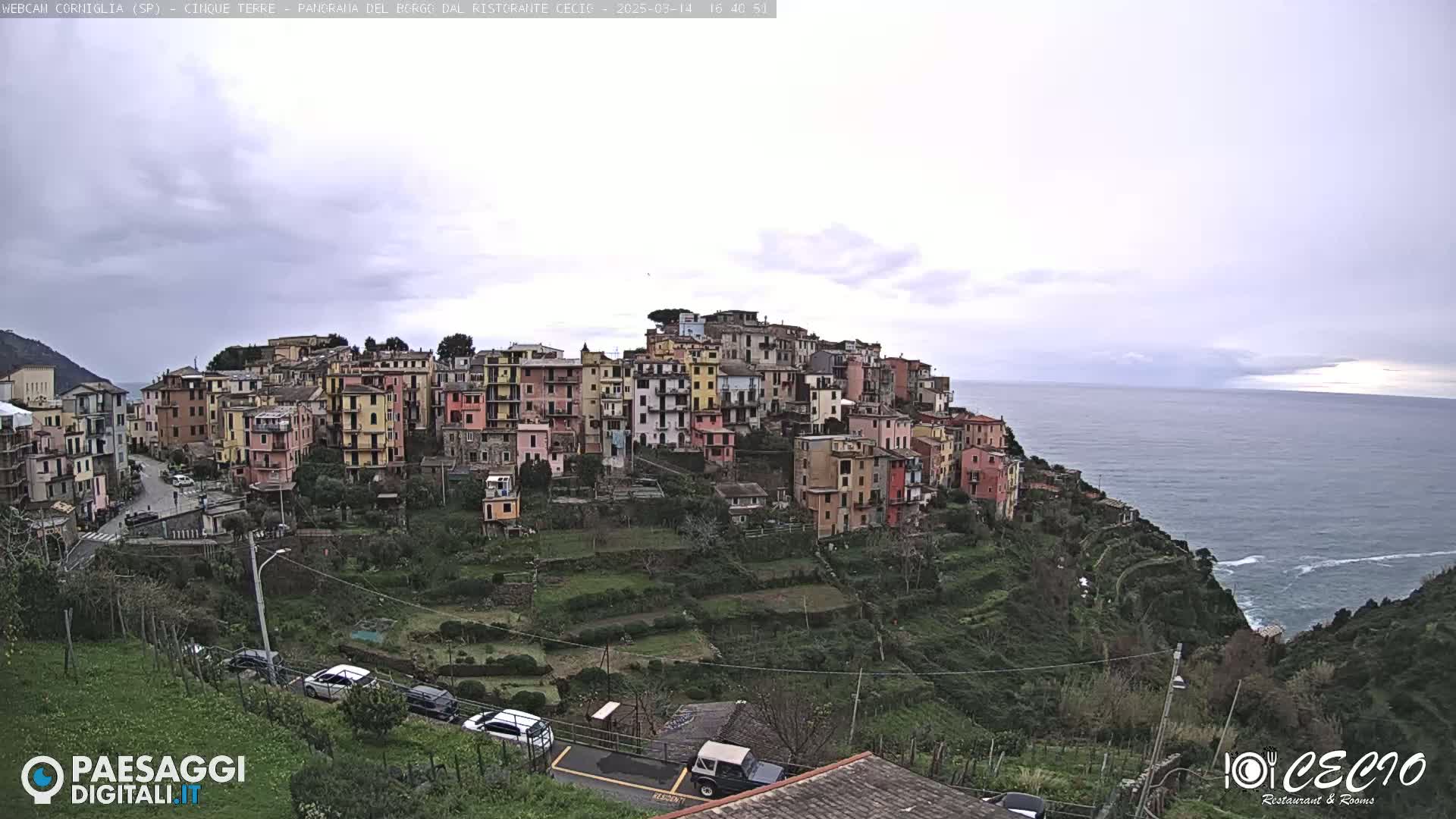 A colorful village clings to a hillside overlooking a calm sea under an overcast sky.