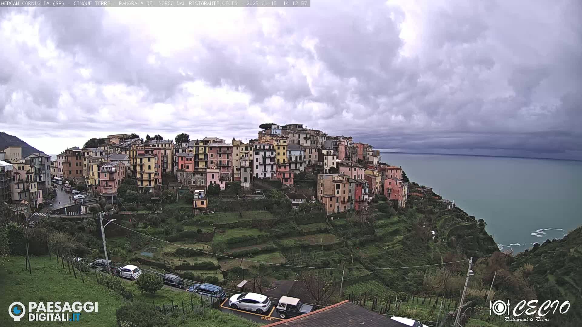 A hillside village of colorful buildings overlooks a teal sea under a cloudy sky.