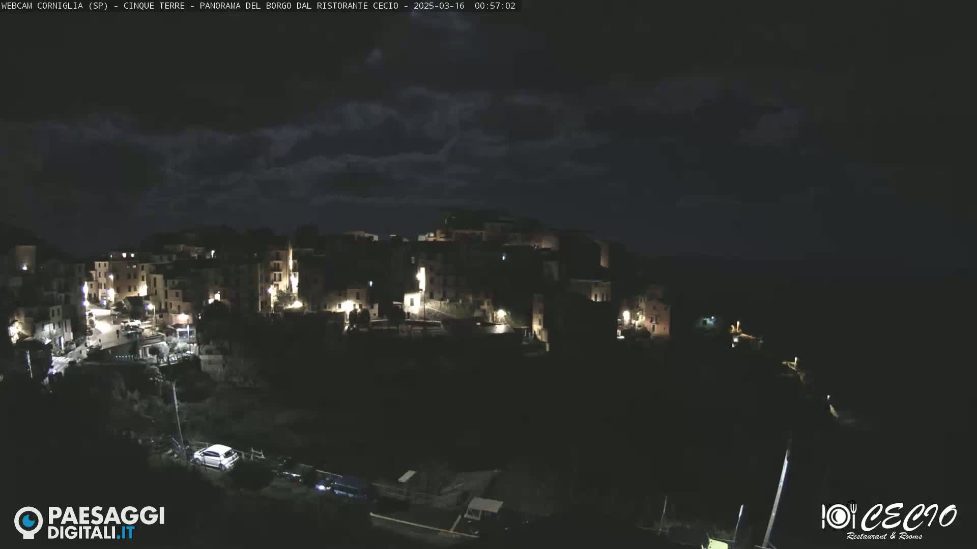 A nighttime view of a small, hilltop town with scattered lights under a dark, cloudy sky.