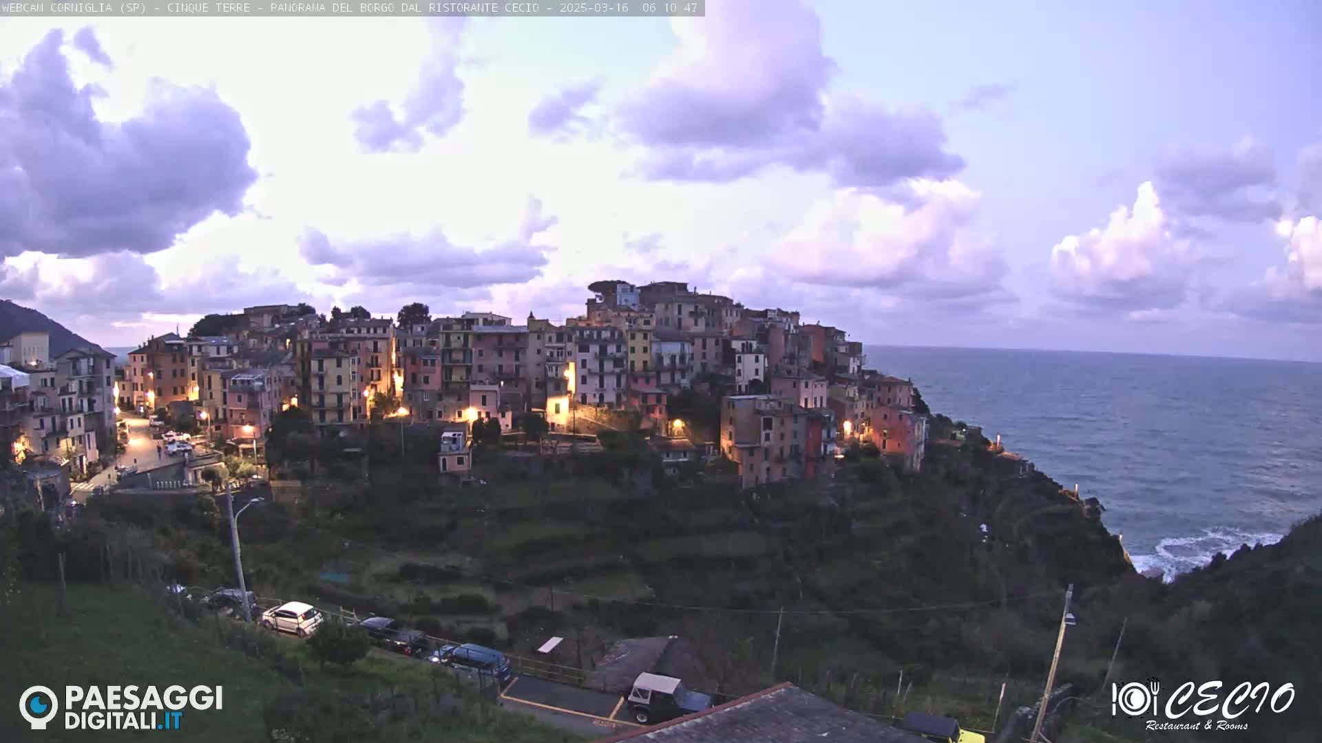 A brightly lit coastal town nestled on a hillside overlooking a calm sea under a partly cloudy twilight sky.
