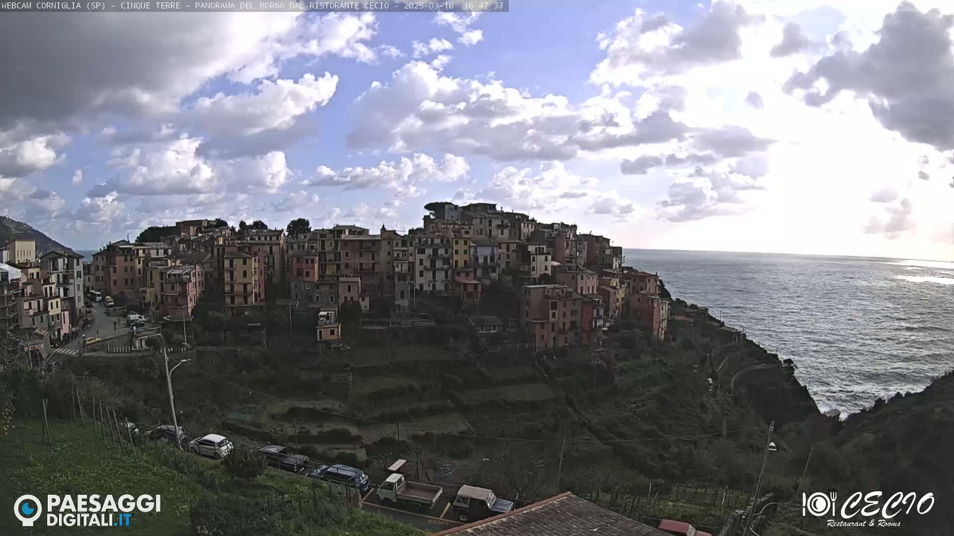 A coastal village of colorful buildings clings to a hillside overlooking a partly cloudy ocean.