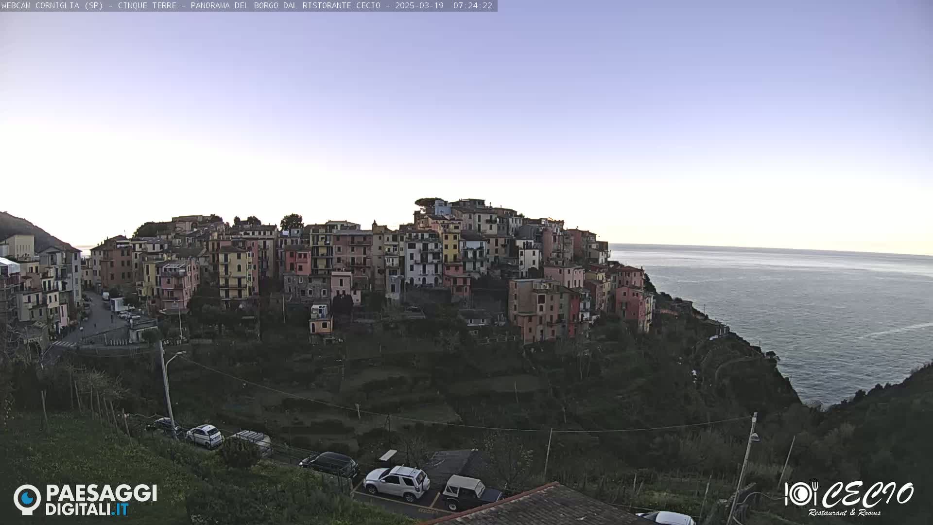 A coastal village of colorful buildings sits on a hillside overlooking a calm sea under a clear, twilight sky.