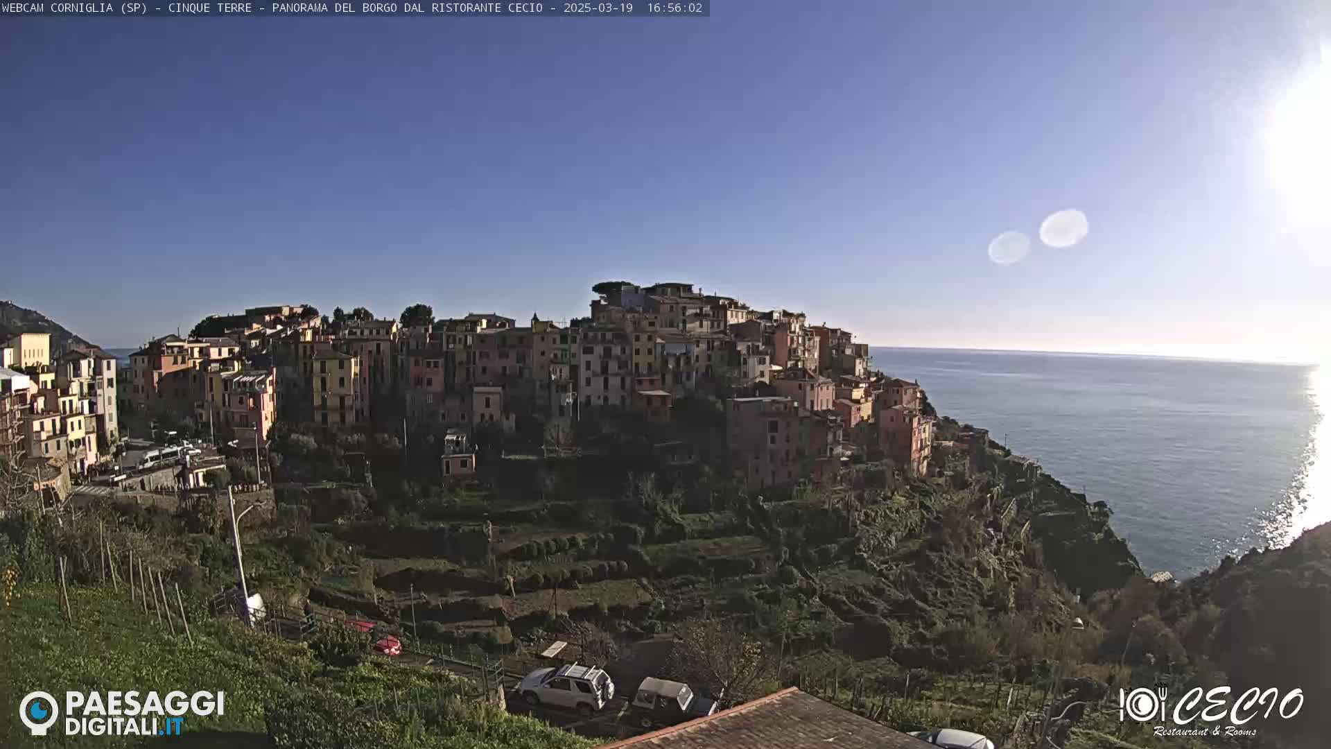 A colorful village clings to a hillside overlooking a calm, blue sea under a clear, sunny sky.