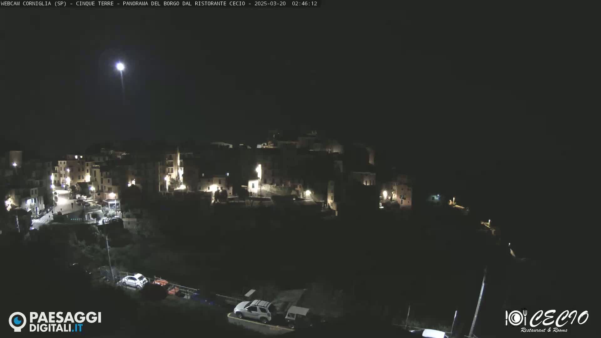 A nighttime, aerial view of a small, illuminated town nestled on a hillside under a clear, dark sky with a bright light visible in the distance.
