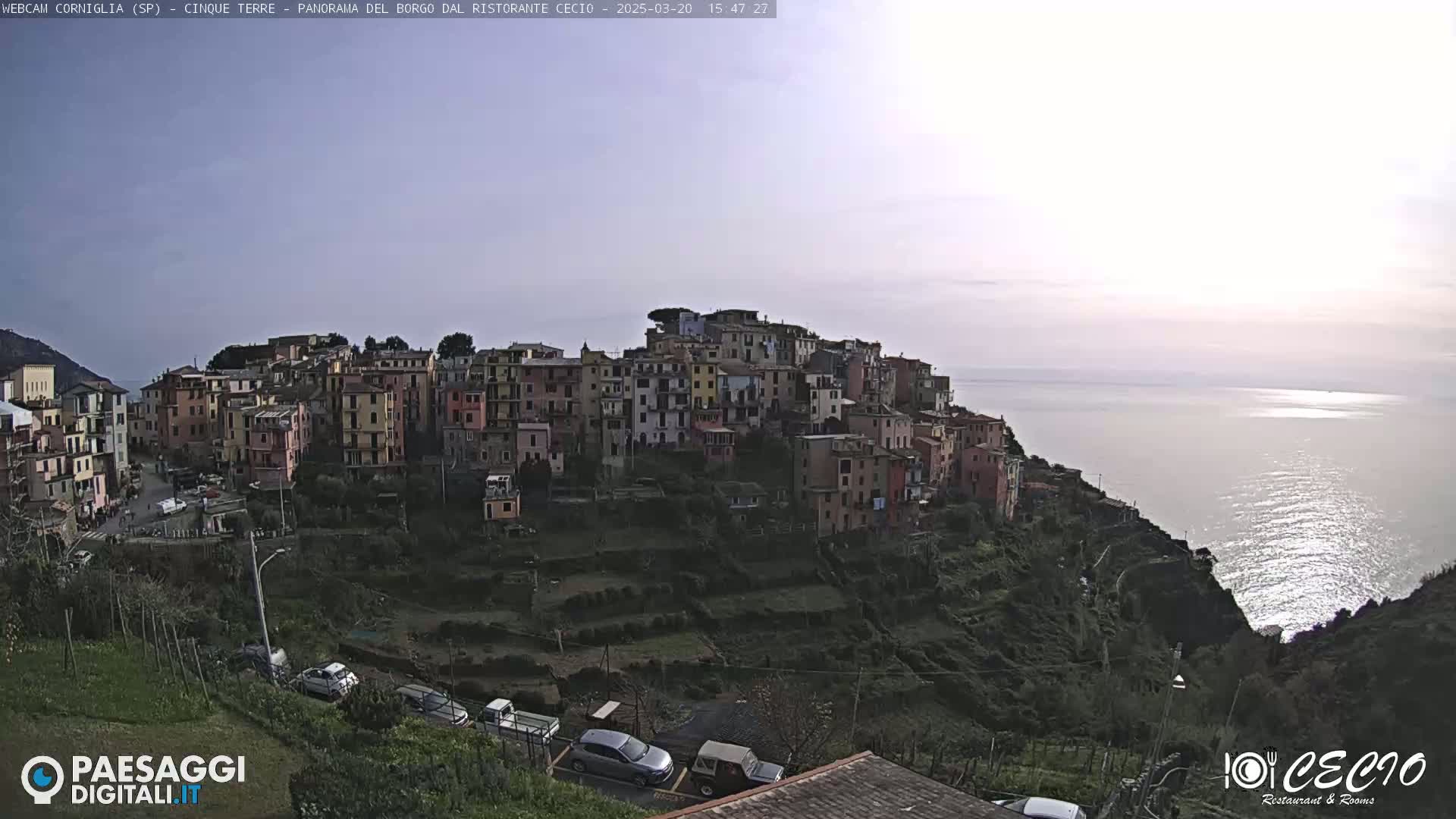 A hillside village of colorful buildings overlooks a calm sea under a mostly clear, light sky.