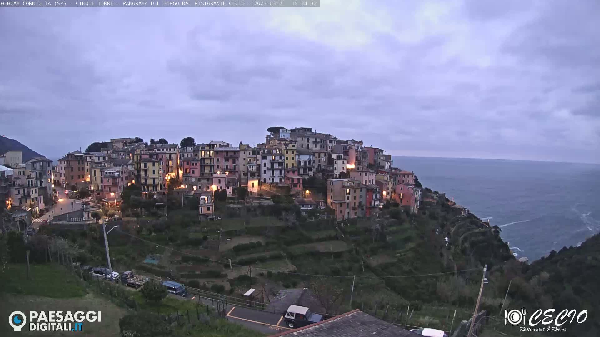 A hilltop village with illuminated buildings overlooks a calm, dark ocean under a twilight sky.