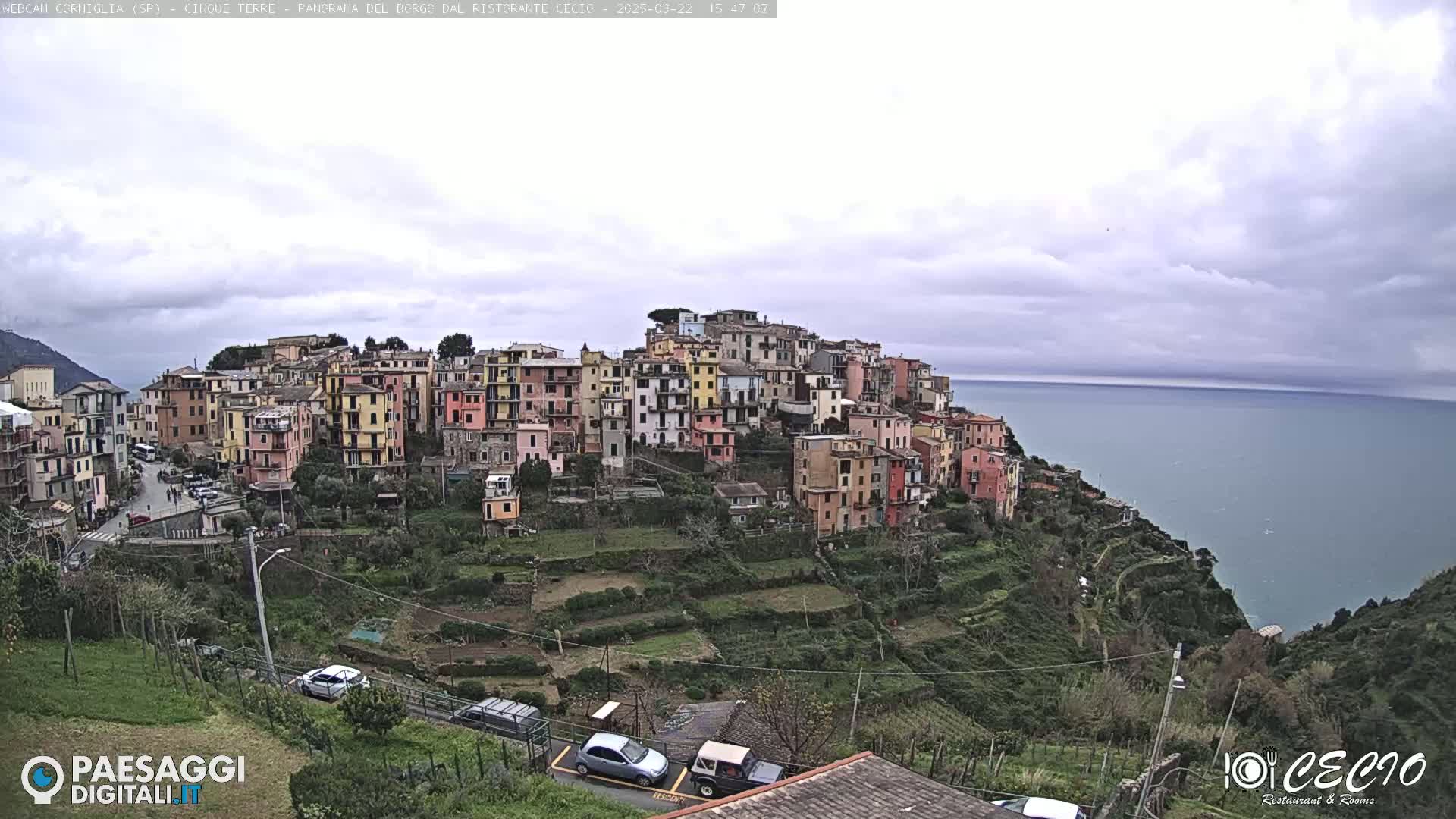 A hillside village of colorful buildings overlooks a calm sea under a mostly cloudy sky.