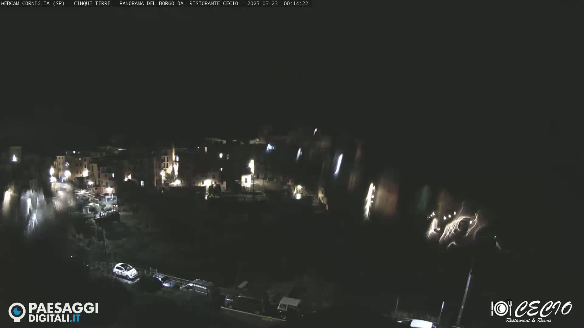 A nighttime view of a small, illuminated town nestled into a hillside, with a few cars parked below.
