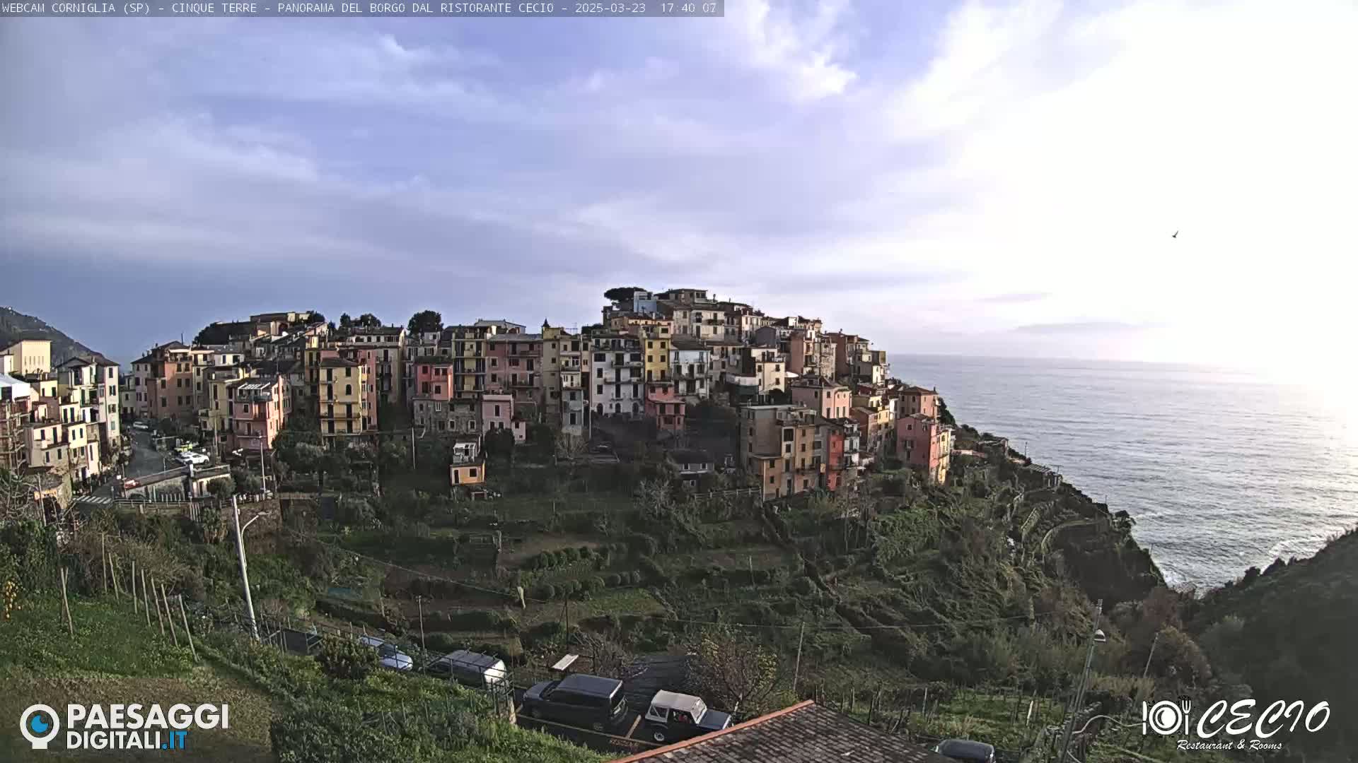 A colorful village clings to a hillside overlooking a calm ocean under a partly cloudy sky.