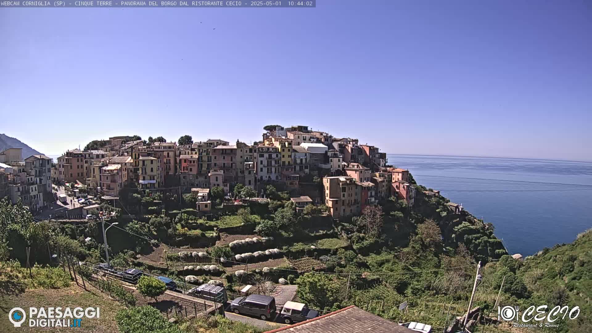 A colorful village clings to a hillside overlooking a calm blue sea under a clear blue sky.