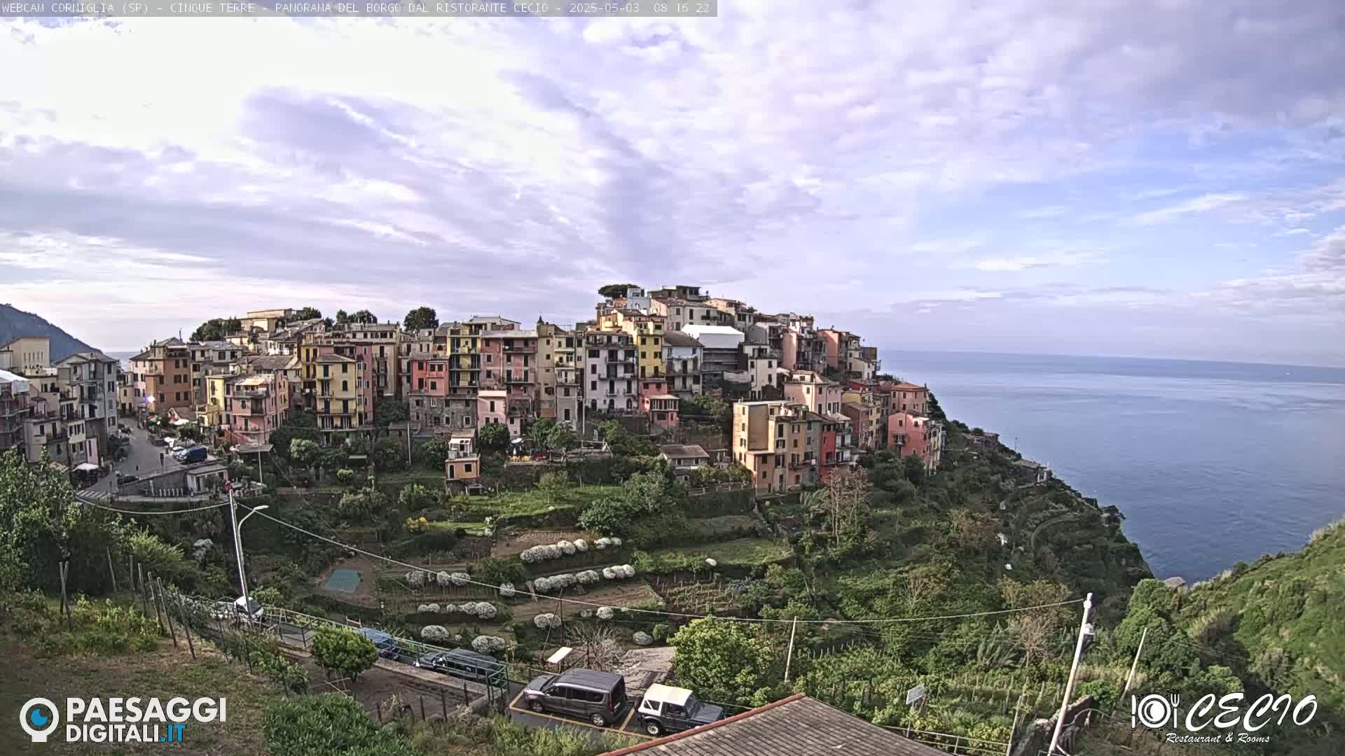 A colorful village clings to a hillside overlooking a calm ocean under a mostly cloudy sky.