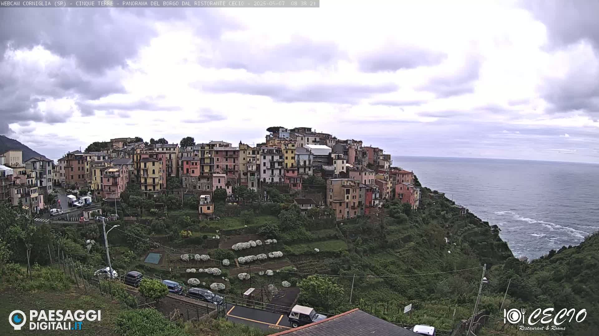 A hillside village of colorful buildings overlooks a calm ocean under a cloudy sky.