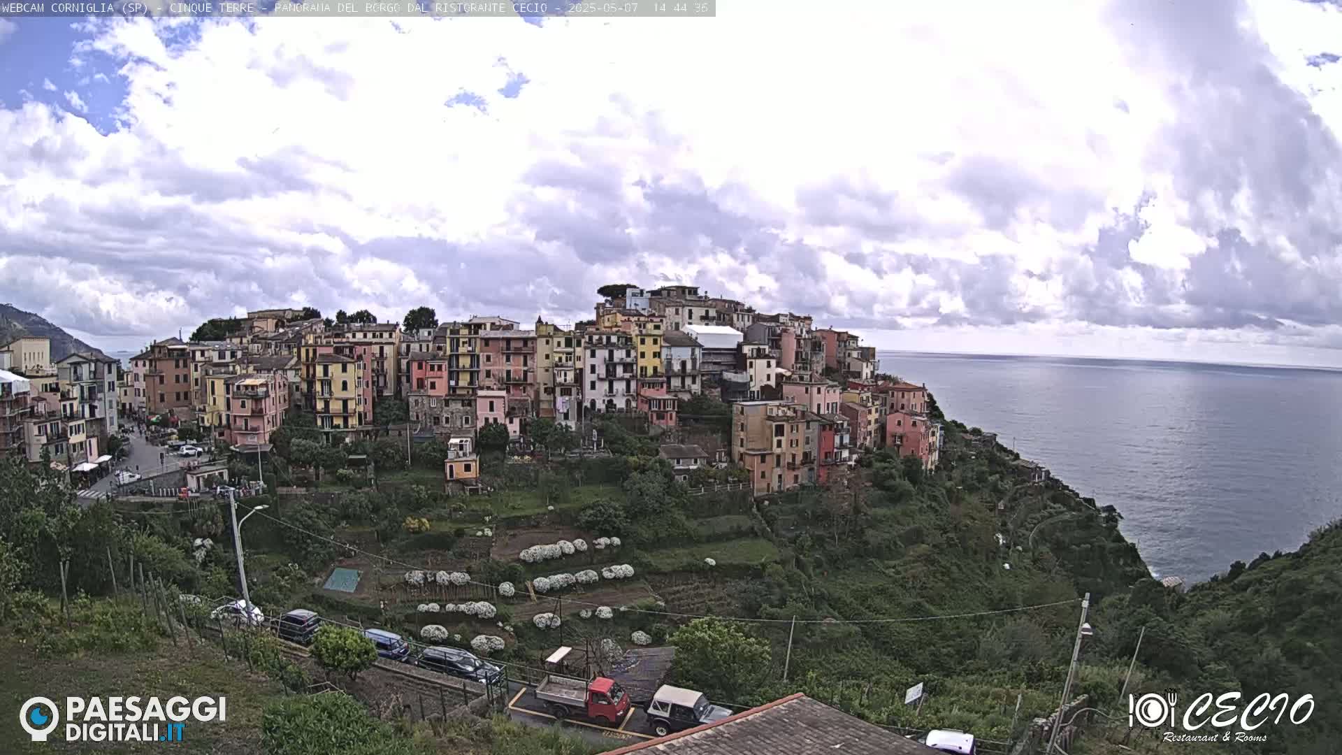 A colorful village clings to a hillside overlooking a calm sea under a partly cloudy sky.