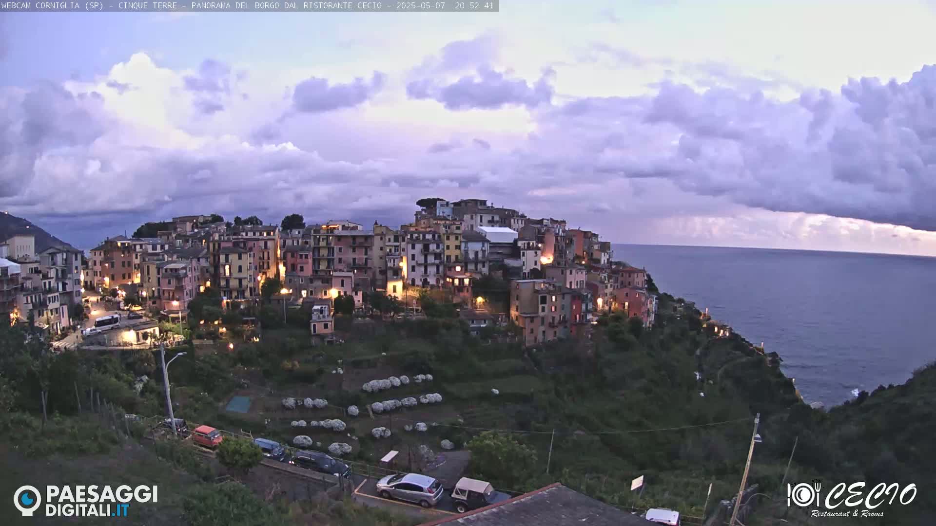A coastal town with illuminated buildings sits on a hillside overlooking a calm sea under a partly cloudy twilight sky.