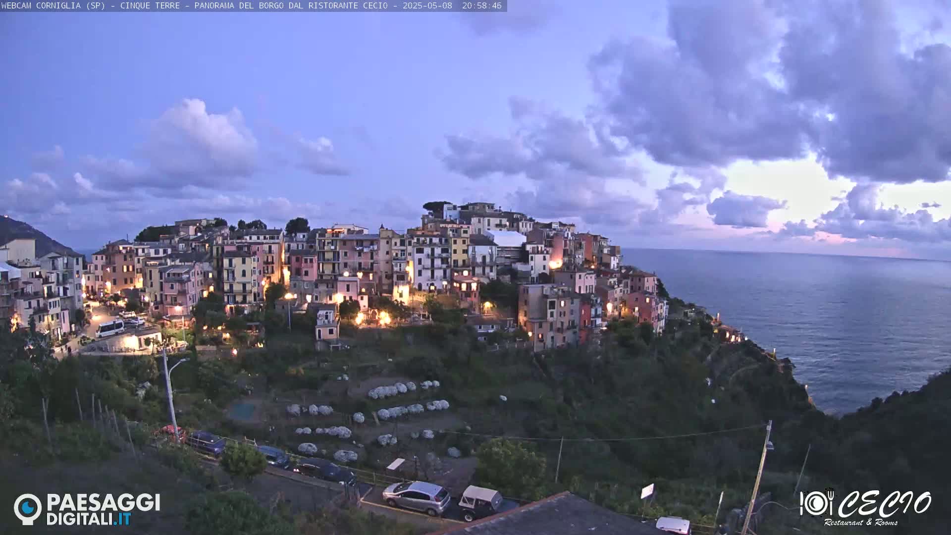 A brightly lit village perched on a hillside overlooking a calm sea under a partly cloudy twilight sky.